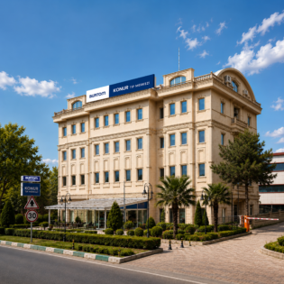 Beige multi-story building with blue sign reading 'BURTON TIP MERKEZI' and 'KONUR', glass entrance, and landscaped forecourt under a blue sky.