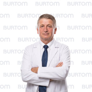 Middle-aged male doctor in a white lab coat with a navy tie stands with arms crossed, looking at the camera.